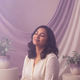 Portrait of a smiling South Asian woman, late 20s, against a soft lavender and warm cream backdrop.
